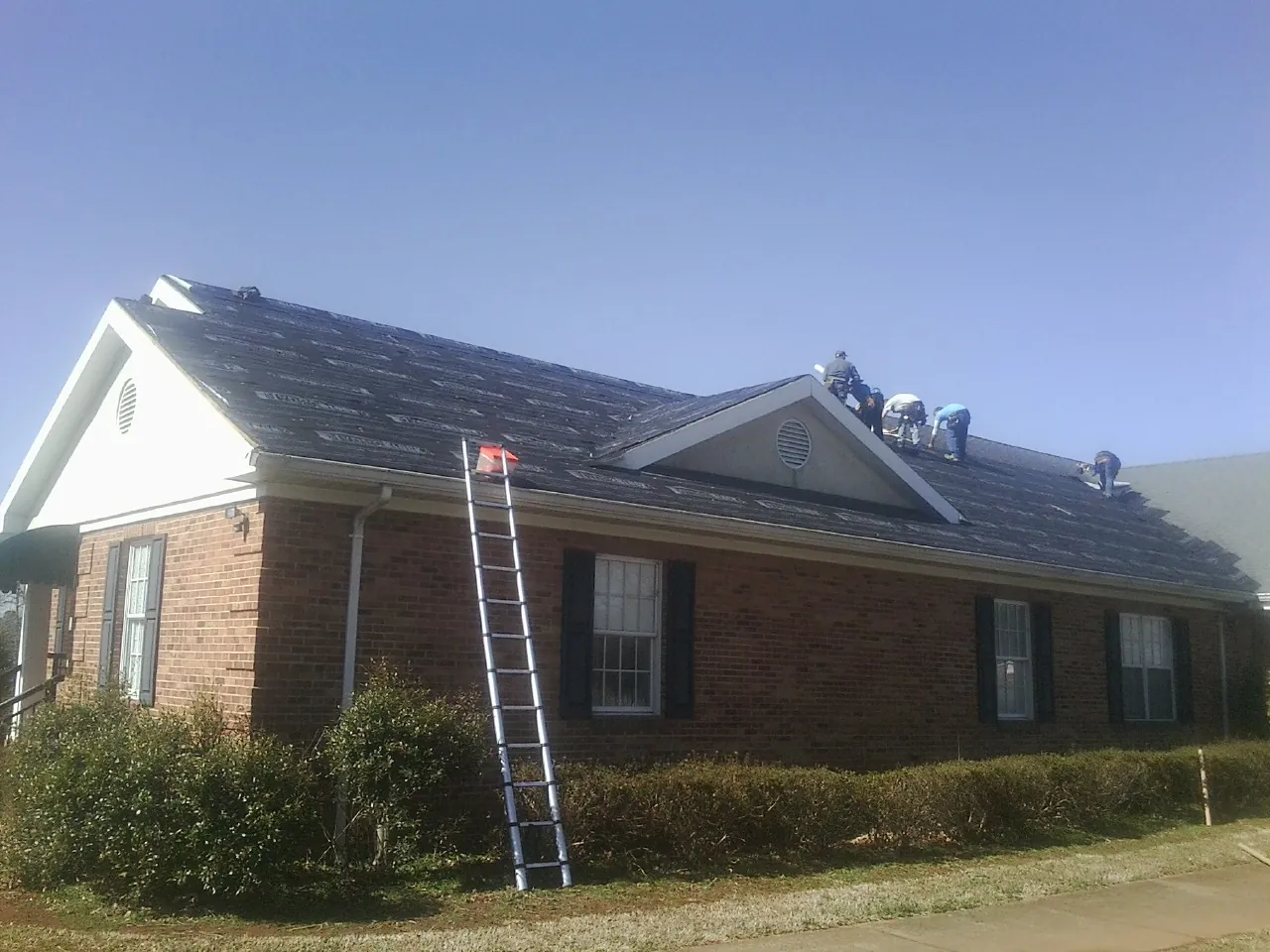 Crew tearing shingles off a brick ranch roof with a ladder set — Metal Roof Repair work by our crew in Sylva