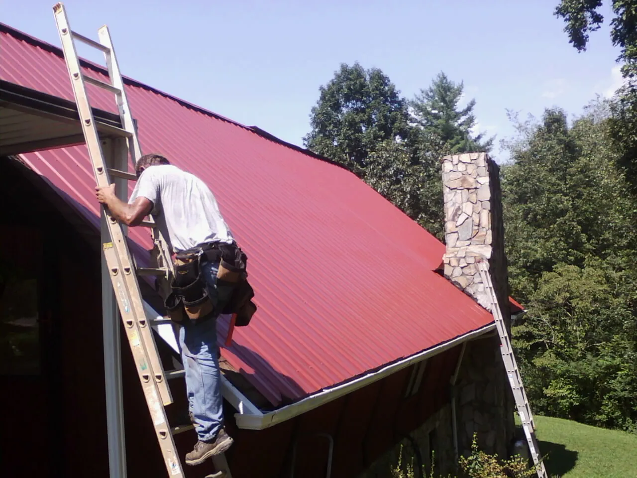 Roofer on a ladder beside a red metal roof and stone chimney — Soffit & Fascia Repair by Roofing Brothers in Sylva